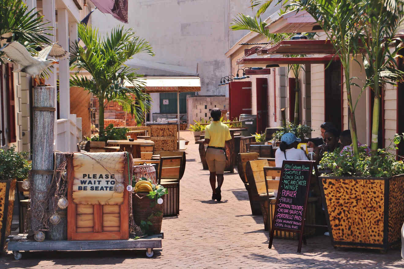 Quaint outdoor dining area in Nassau with wooden tables and chairs, surrounded by lush potted plants and a "Please Wait to Be Seated" sign, with a staff member in a yellow shirt walking through