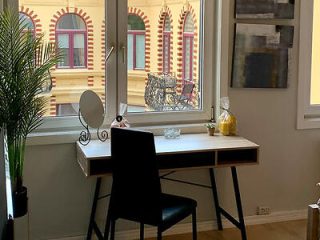 A cozy workspace by a window with a view of European-style architecture, featuring a white modern desk, black chair, and decorative mirror, complemented by indoor greenery.