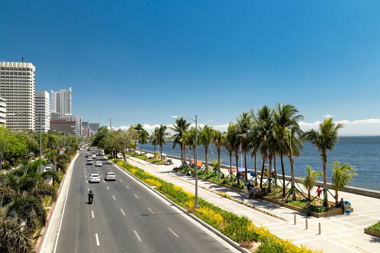 Sunny view of Roxas Boulevard in Manila, with palm trees lining the bay walk, against a backdrop of high-rise buildings and a clear sky