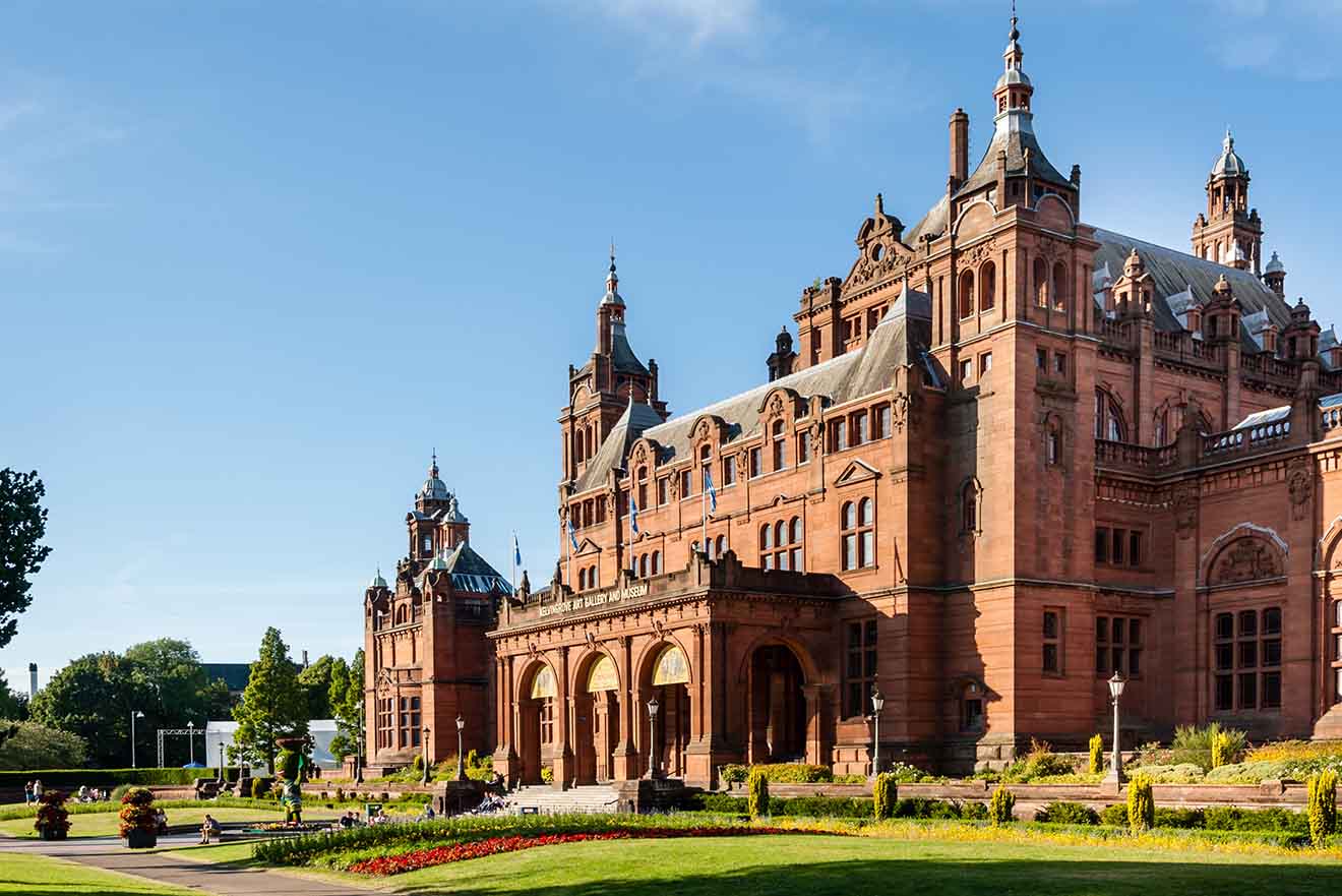 The distinguished Kelvingrove Art Gallery and Museum in Glasgow, presented in warm sandstone against a blue sky, with people enjoying the manicured lawns