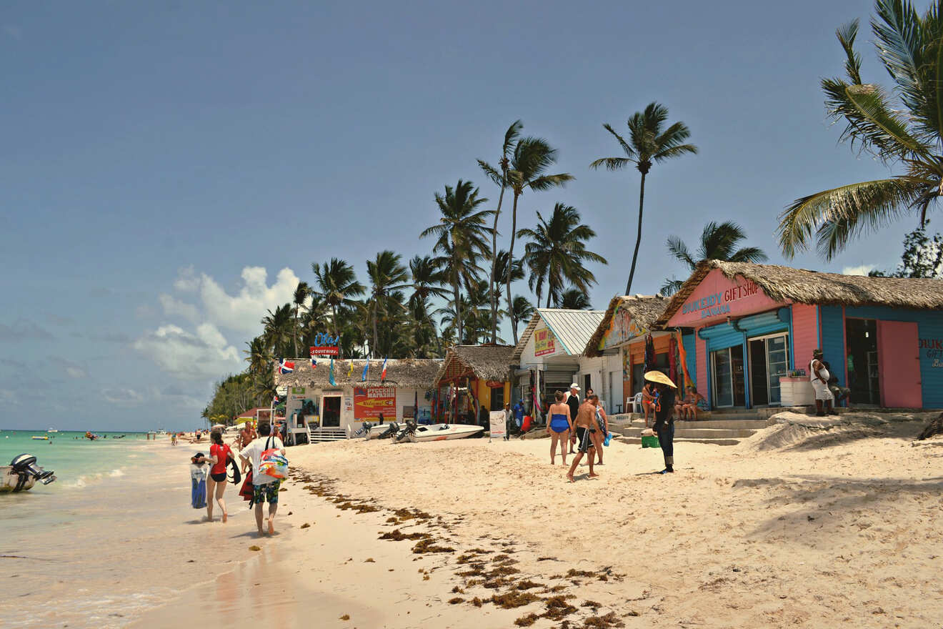 Beachfront scene with visitors walking by colorful tropical storefronts under coconut palms on a sandy beach.
