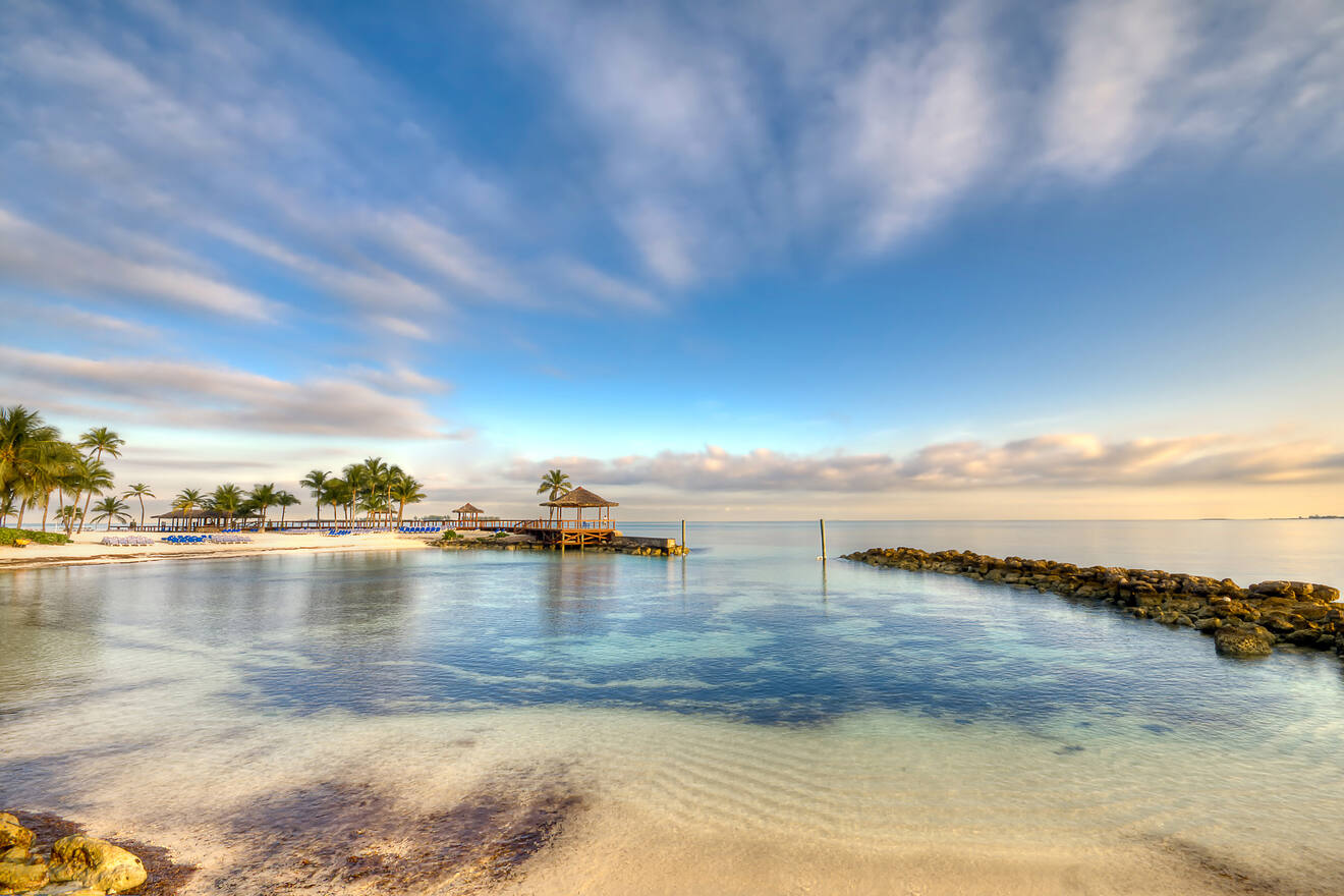 Tranquil beachfront in Nassau during sunrise or sunset with a gazebo, palm trees, and calm waters reflecting the soft pastel colors of the sky.