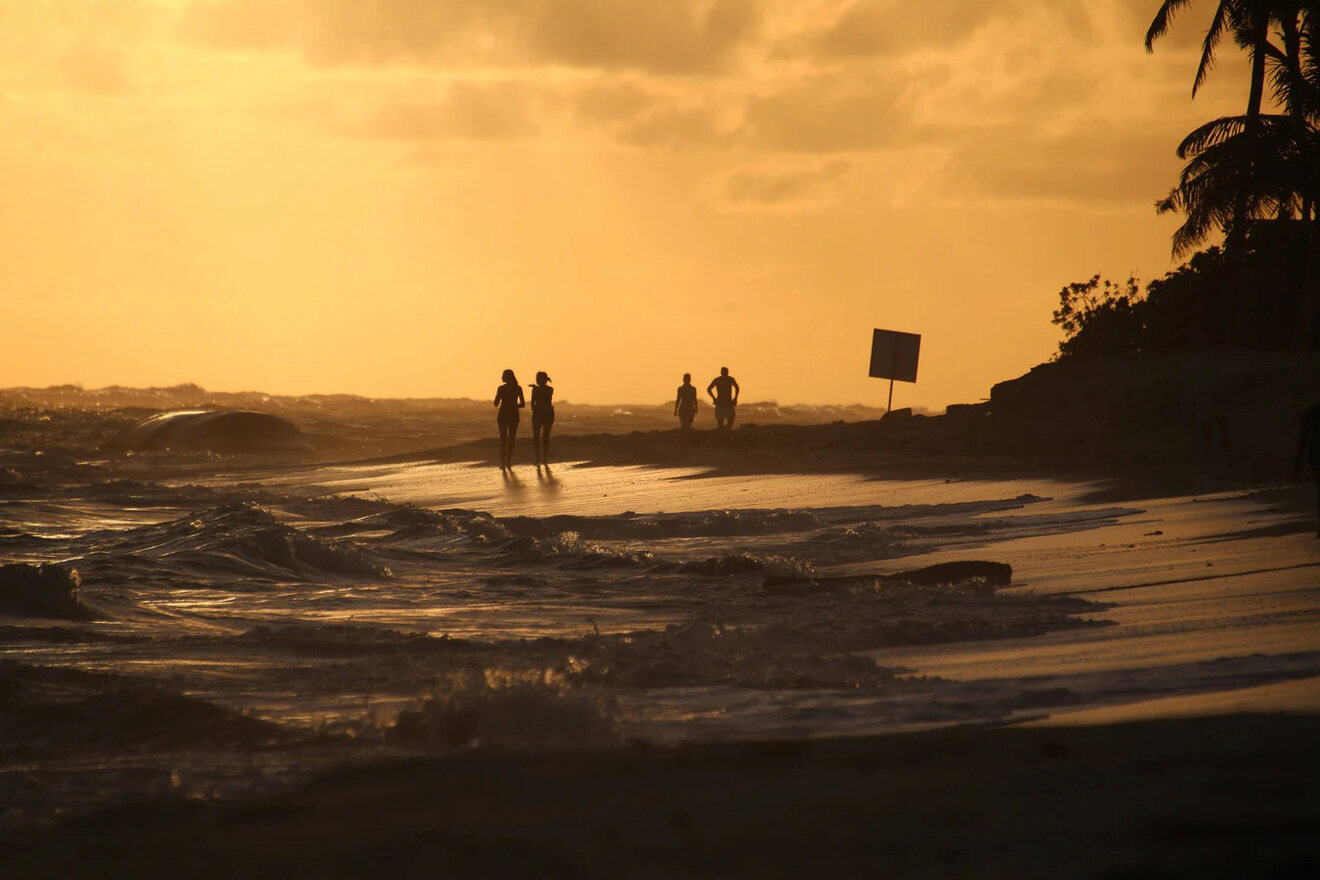 Silhouetted figures on a beach at sunset, with waves lapping the shore and a darkening sky, creating a tranquil end-of-day scene.
