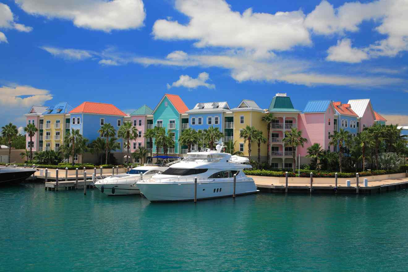 Colorful waterfront row of pastel-colored houses in Nassau with a luxury yacht moored in the calm turquoise waters, under a partly cloudy sky.