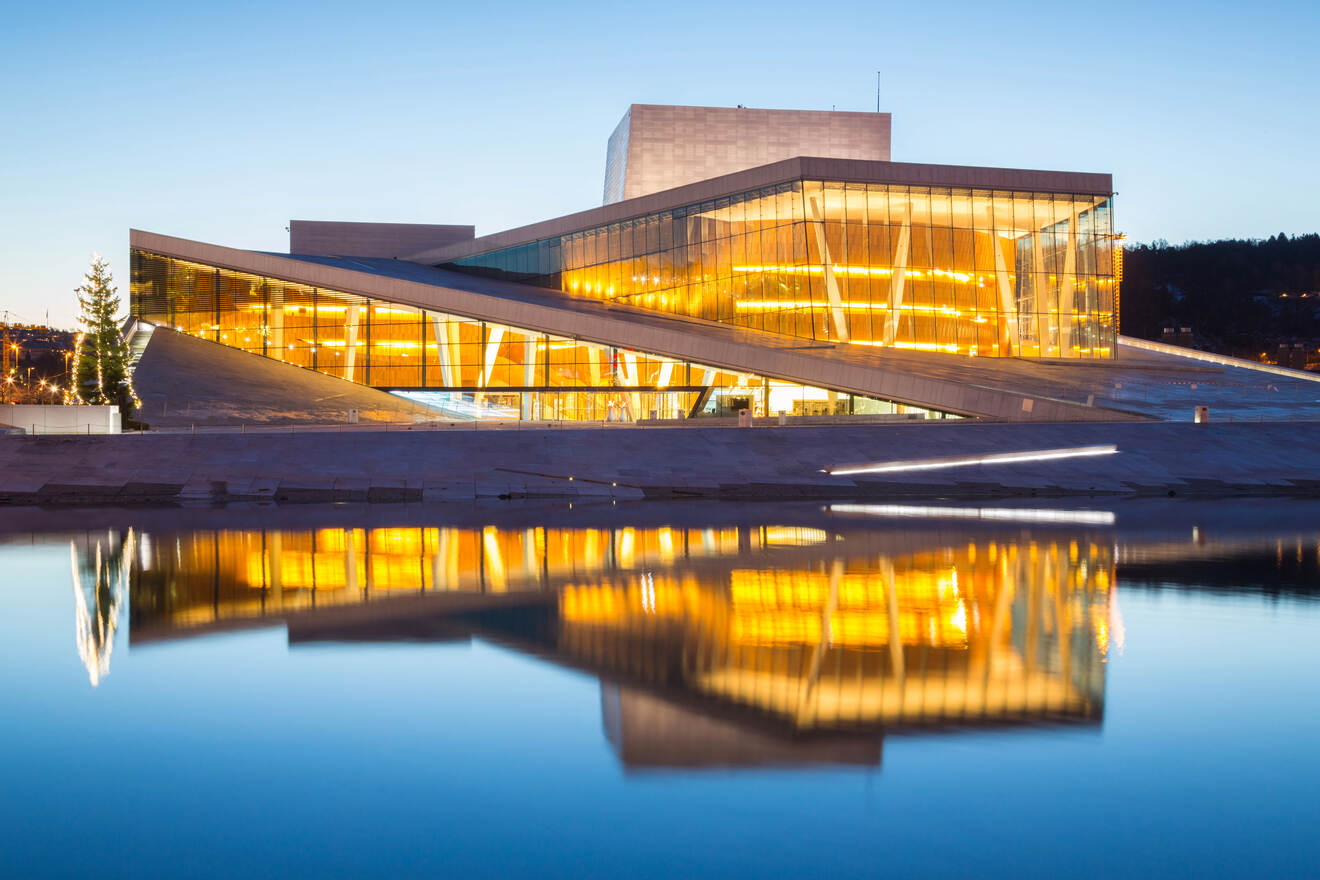 The Oslo Opera House at twilight, its contemporary architecture illuminated and reflecting in the calm waters in front of it.
