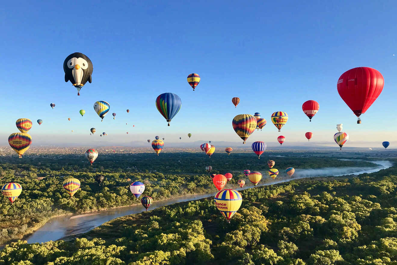 Hot air balloons in various colors and designs float over a lush green landscape with a winding river beneath them under a clear blue sky.