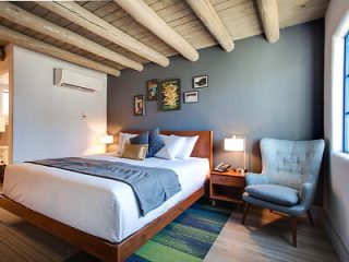 A modern bedroom with a wooden ceiling, double bed with gray bedding, bedside tables with lamps, framed pictures on a blue wall, and a gray upholstered chair.