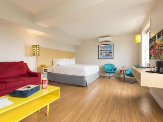 Modern hotel room with a minimalist design featuring a red sofa, colorful chairs, and a large bed with a bright window view