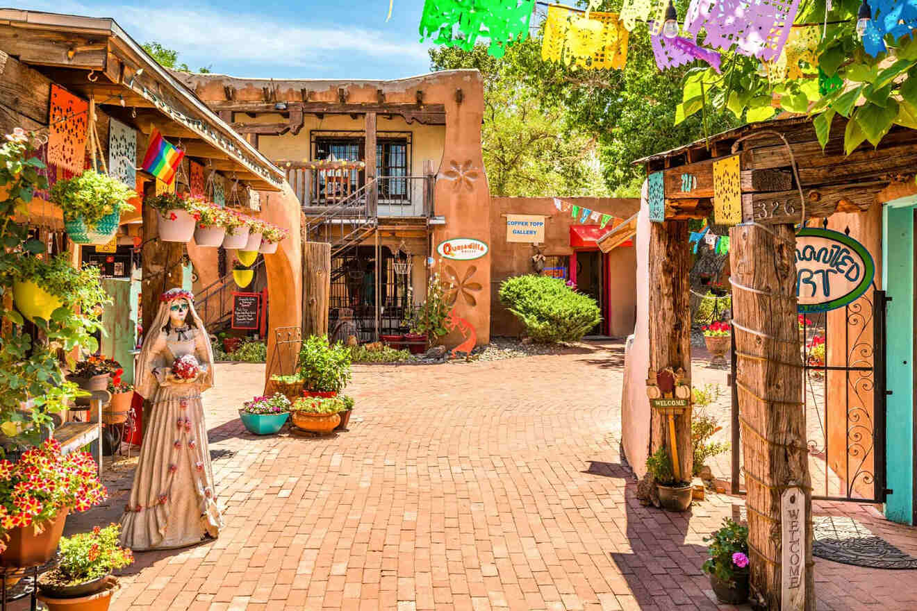 A vibrant, colorful courtyard is adorned with plants, flowers, and festive decorations. Signs for shops like an art gallery and café are visible. The buildings feature rustic, Southwestern architecture.