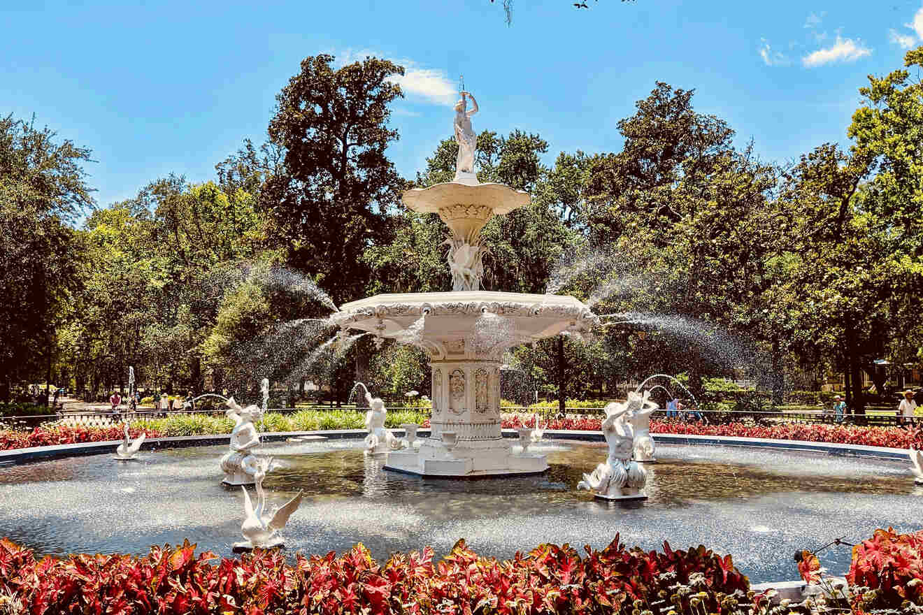 Forsyth Park's iconic white fountain surrounded by red flowers and lush greenery under a sunny sky in Savannah, Georgia.