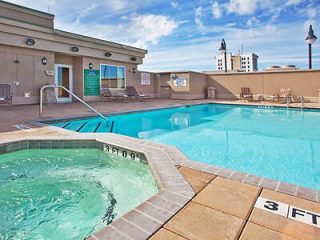 Hotel rooftop pool and hot tub area with lounge chairs and a cityscape in the background on a clear day.