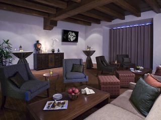 A cozy lounge with armchairs, a sofa, and tables. A television is mounted on the wall, and the room features potted plants, dim lighting, and a wooden ceiling.