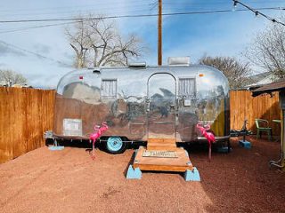 A silver Airstream trailer with pink flamingo lawn ornaments standing on reddish gravel, set against a wooden fence background with bare trees and a cloudy sky.