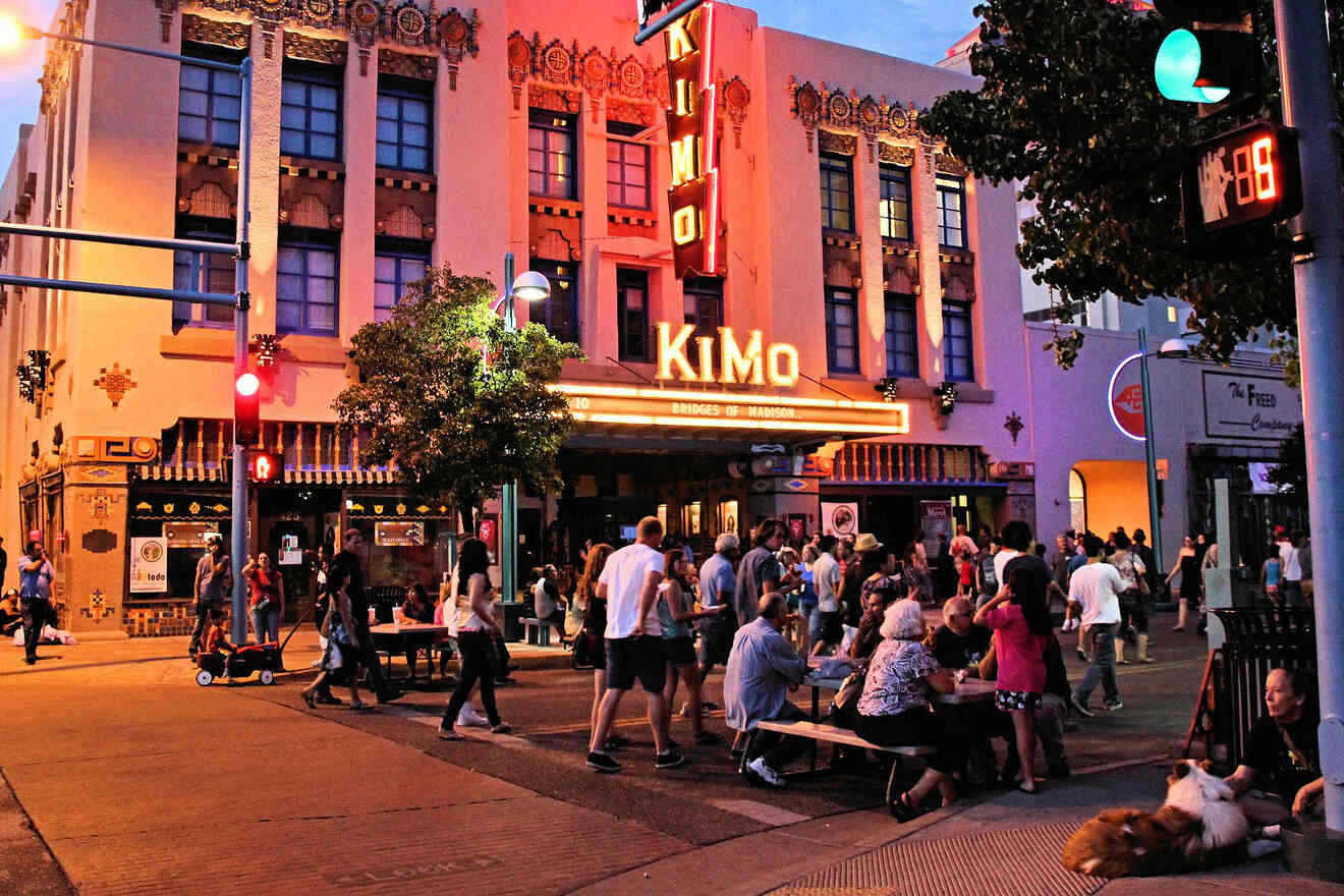 People gather outside the illuminated KiMo Theatre at dusk, with some sitting at tables and others walking. The vibrant street scene includes a traffic light, crosswalk, and nearby storefronts.