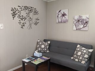 A small waiting room with a gray sofa, patterned pillows, a coffee table with magazines, tissue box, metal wall art, and two floral pictures.