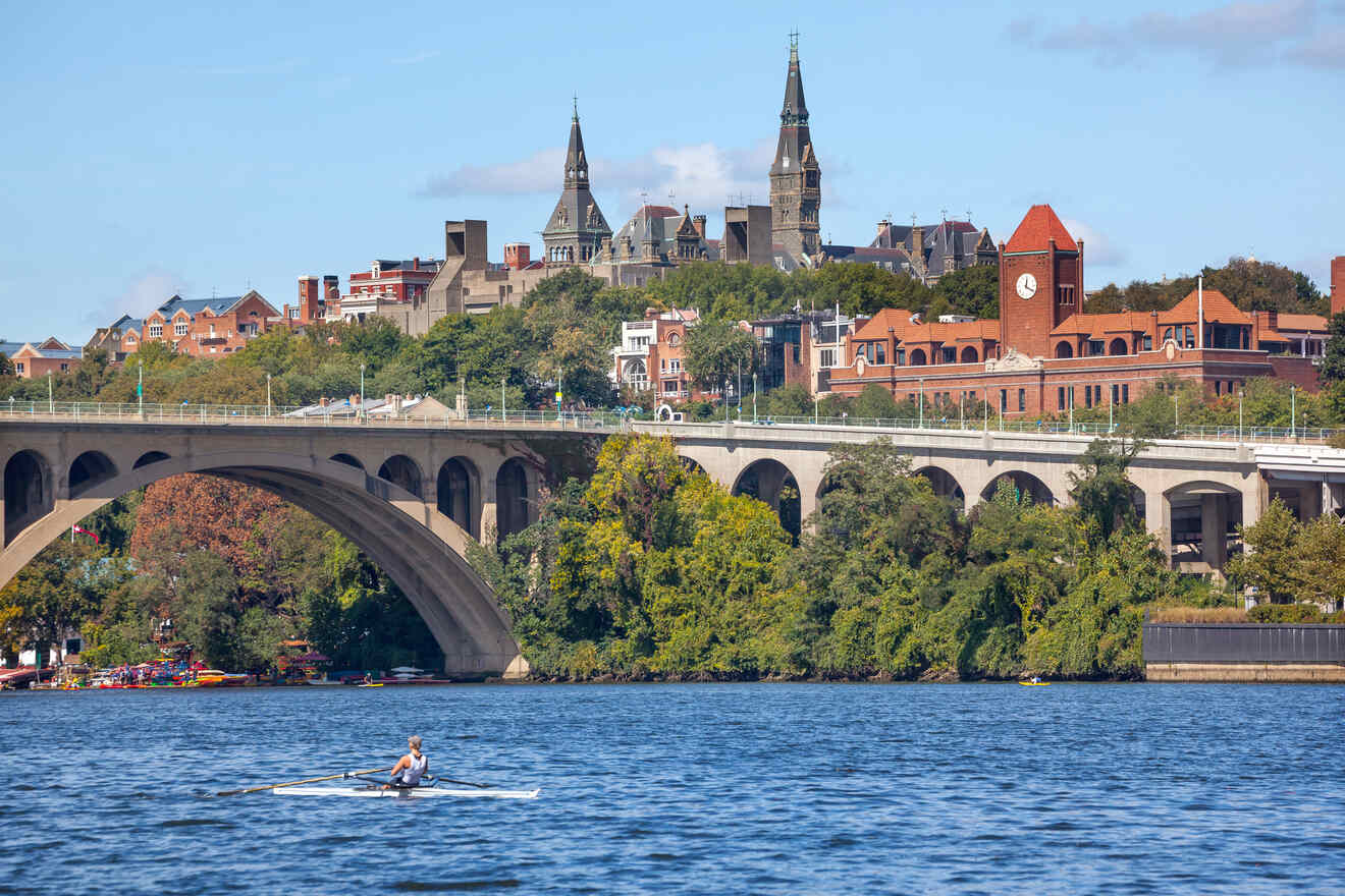 A lone rower gliding on the tranquil waters of the Potomac River with the Georgetown University campus adorning the hilltop in the background.
