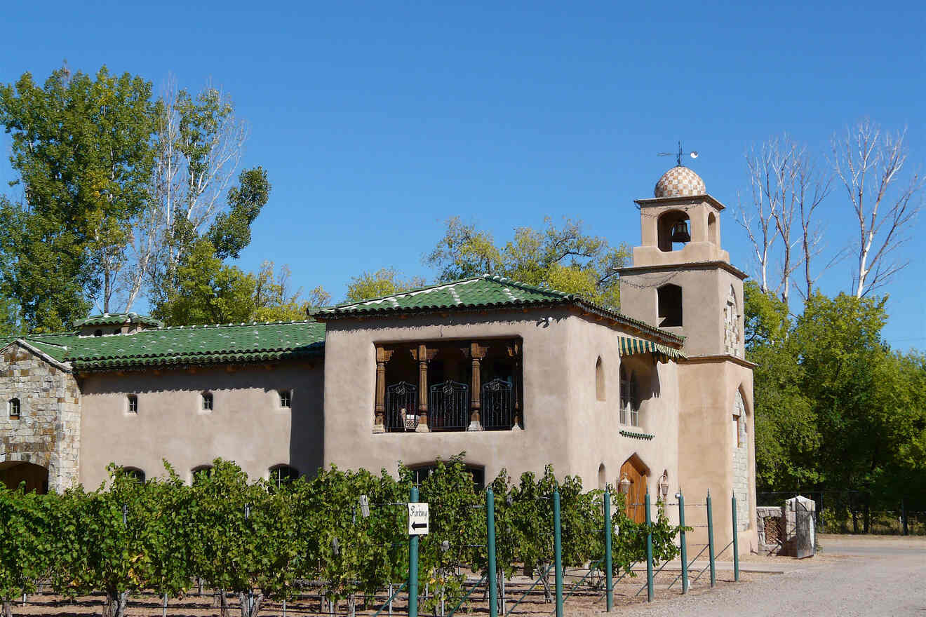 A two-story adobe building with green-tiled roofs and a small tower, surrounded by a vineyard and trees under a clear blue sky.