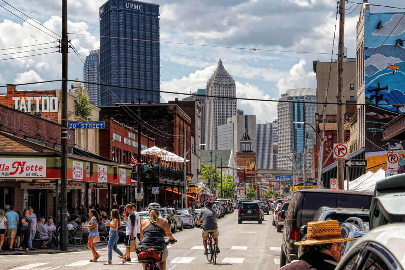 Busy street scene in Pittsburgh with pedestrians, cyclists, and a variety of shops under a clear sky