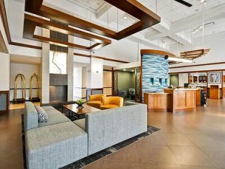 A modern hotel lobby featuring a gray sectional sofa, orange chairs, a check-in desk with a blue wave pattern backdrop, luggage carts to the left, and contemporary decor elements.