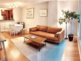 A cozy living room with a large tan sofa, a blue rug, and a wooden coffee table, with a dining area in the background featuring a white table and chairs.