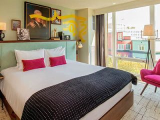 A modern hotel room with a large bed with white linens and black polka dot pattern, accented with red pillows, and a pink chair near a window with city views.