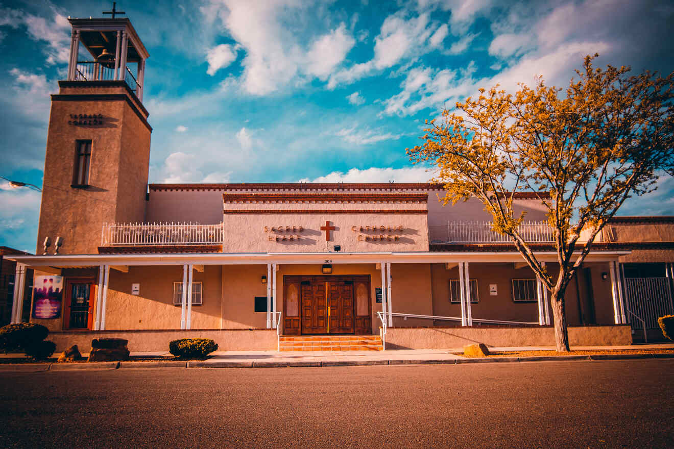 A beige church building with a central wooden door, cross above it, and a bell tower to the left. The facade features a tree on the right and a blue sky with clouds in the background.