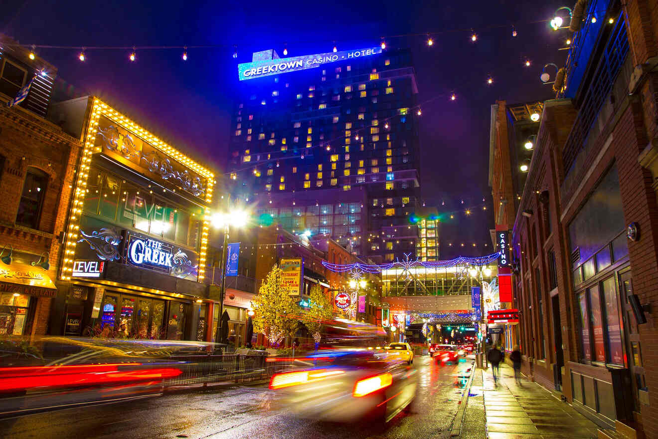 Nighttime street view in Detroit's Greektown showing vibrant neon signs, streaks of traffic lights, and the Greektown Casino Hotel brightly lit in the background