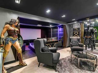 A home cinema room with life-size superhero figures of Wonder Woman and Batman, plush seating, and a contemporary dark-toned decor.