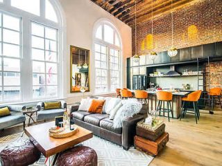 A chic loft-style living space with high arched windows, exposed brick walls, a modern kitchen area, and a cozy lounge setup with a brown sectional sofa.