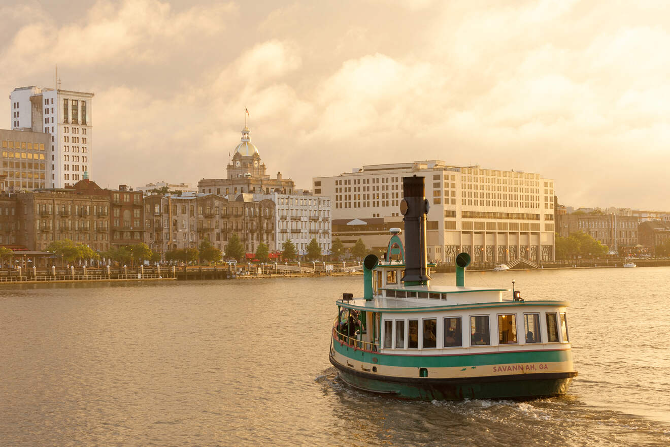 The golden hour sun casts a warm glow over the Savannah River, highlighting a green and white ferry boat marked 'Savannah, GA' as it cruises by a waterfront lined with historic buildings