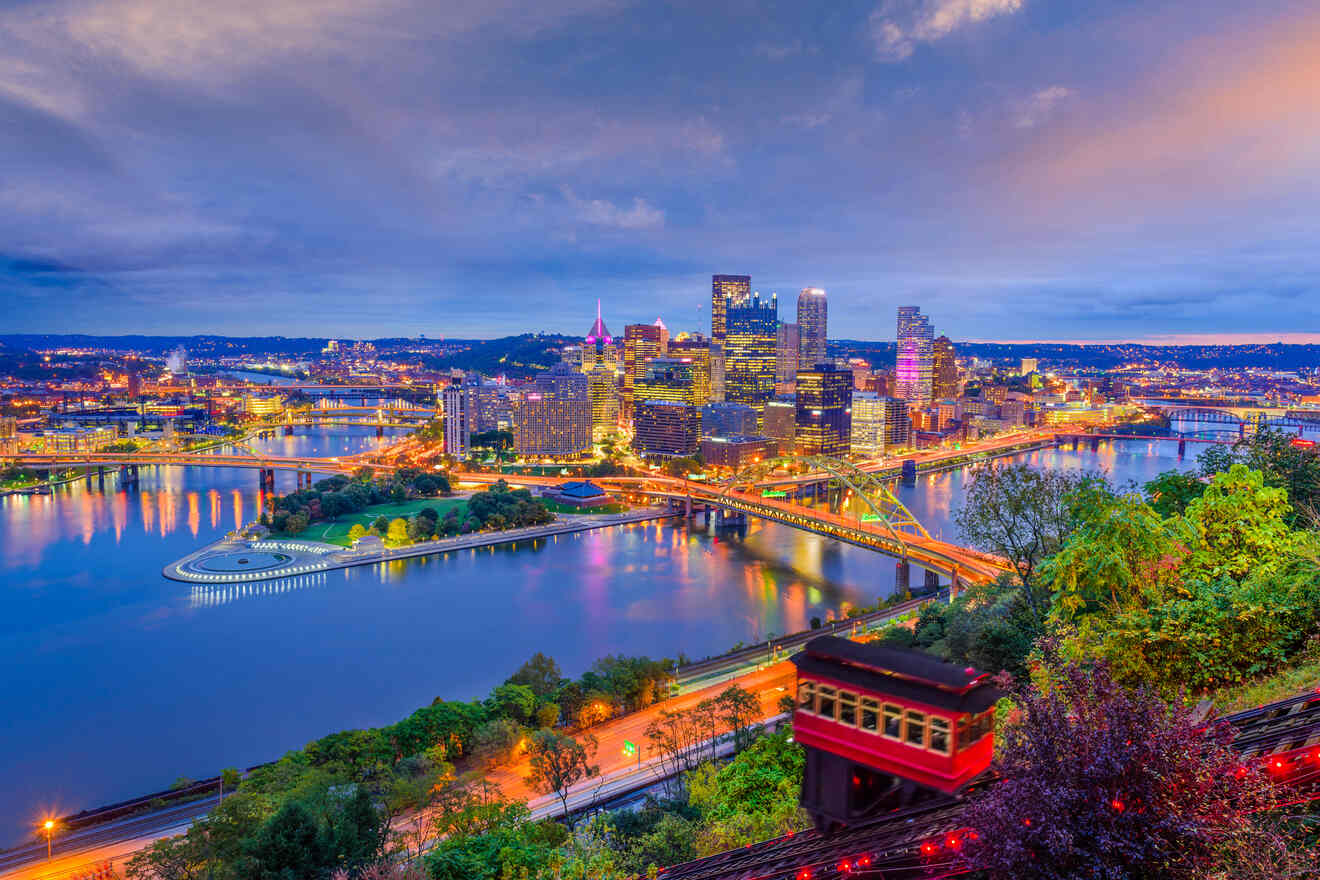 Nighttime view of Pittsburgh skyline showing brightly lit buildings, rivers, and bridges with a scenic overlook