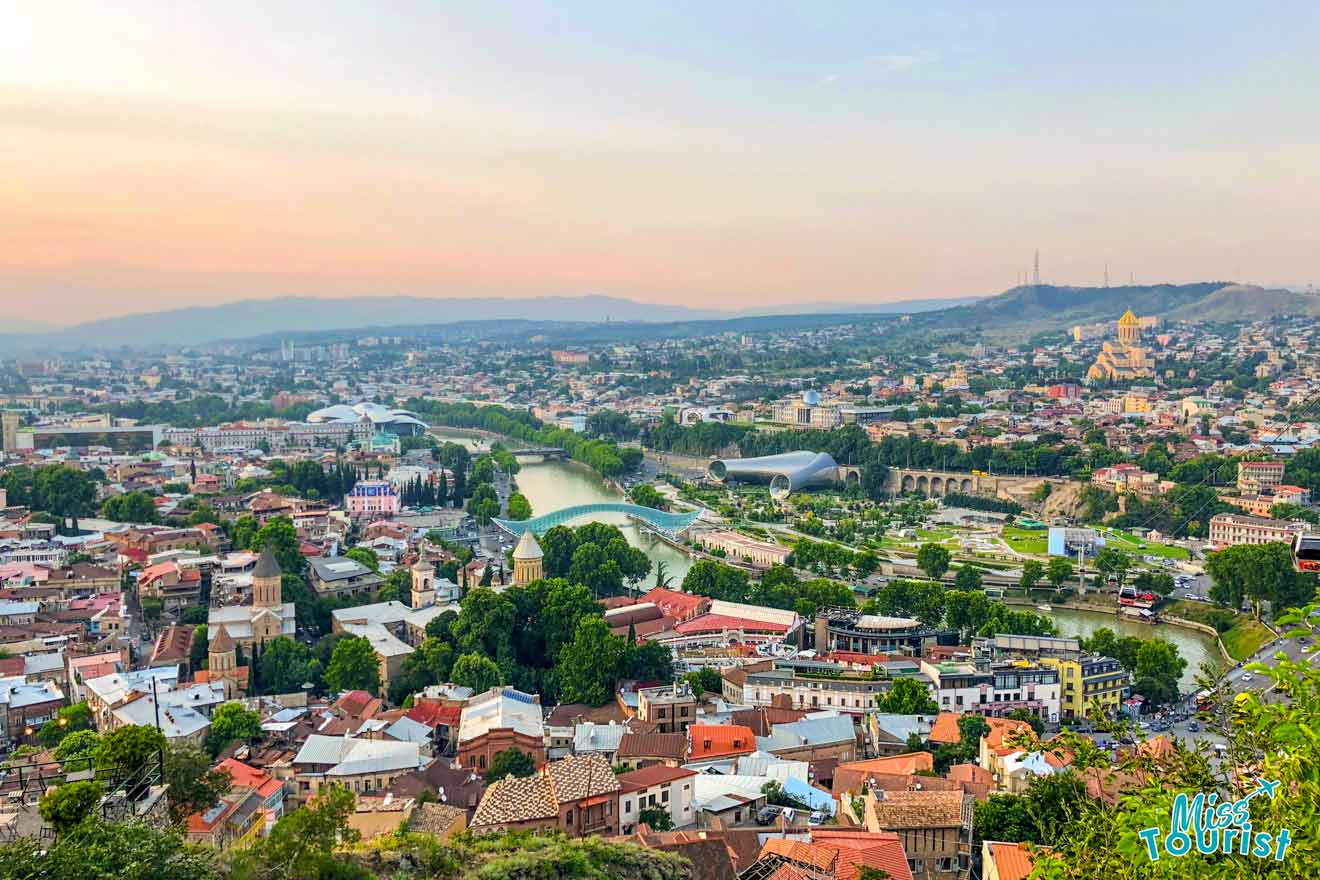 A panoramic view of Tbilisi with the Peace Bridge and Rike Park visible, showcasing the city's blend of historical architecture and modern landmarks under a golden sunset.