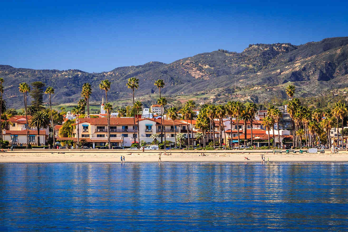 A serene view of Santa Barbara coastline with the calm ocean in the foreground, palm trees, and Spanish-style buildings against the backdrop of the Santa Ynez Mountains under a clear blue sky