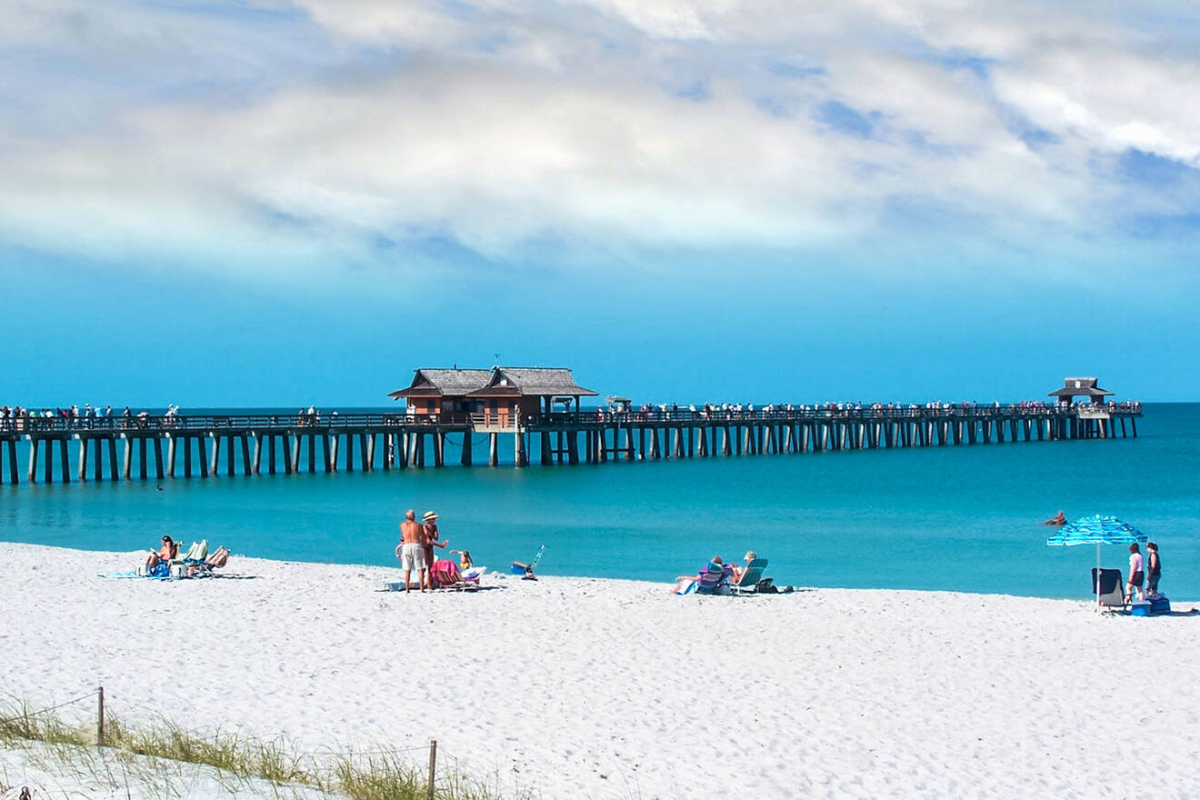 A scenic beach with white sand, people relaxing, a long pier stretching into the ocean, and a clear blue sky.