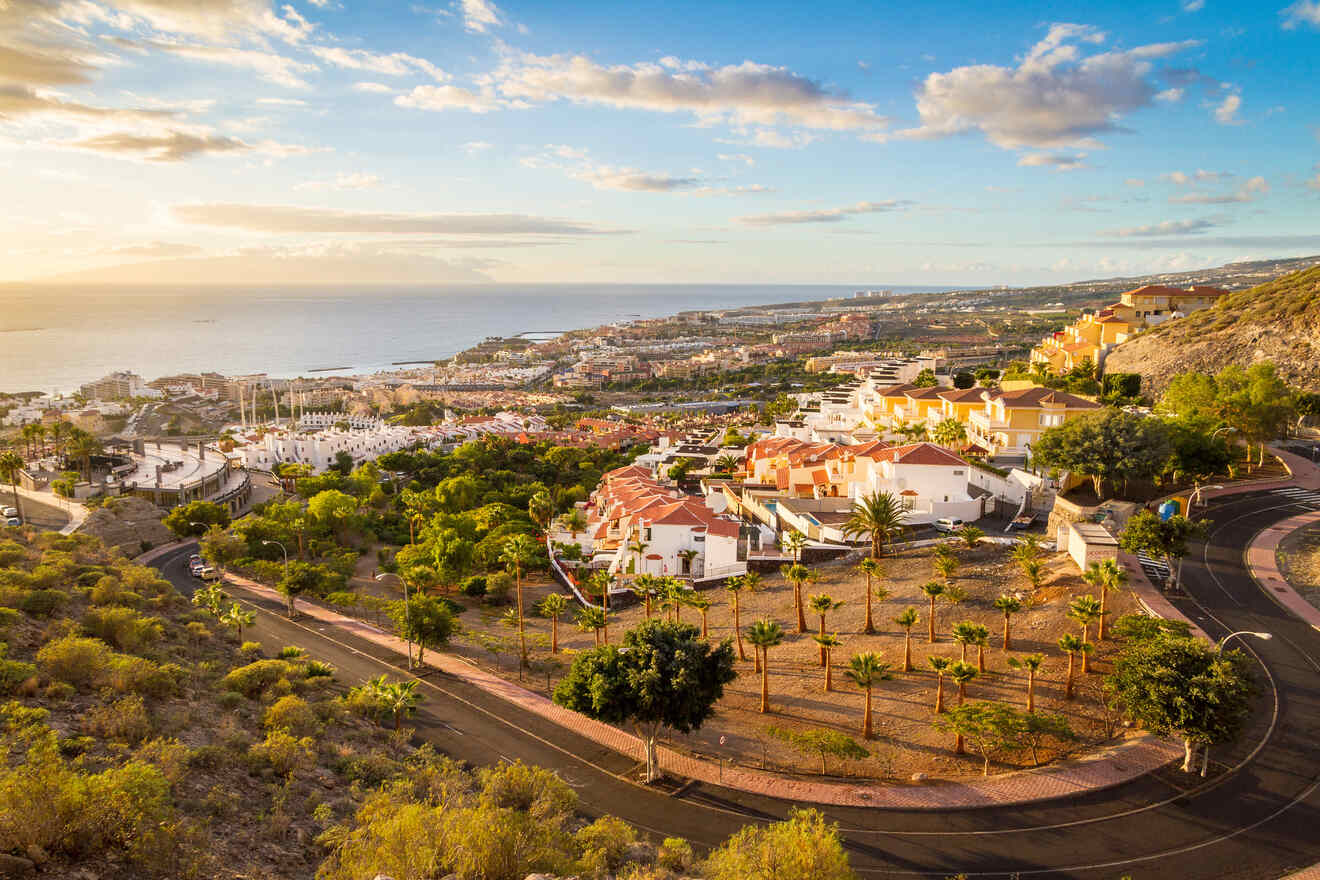 A scenic view of a coastal town in Tenerife with winding roads, white buildings, and lush greenery, overlooking a calm ocean at sunset.