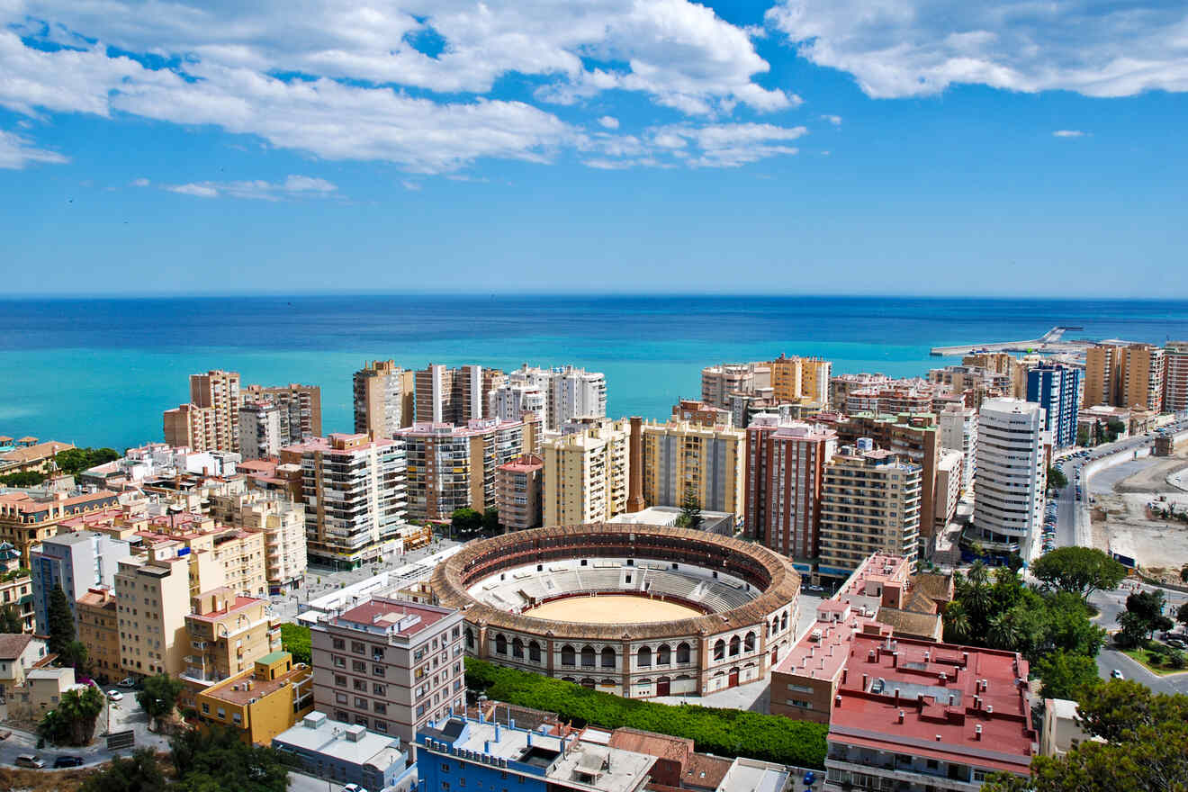 Aerial view of Malaga with a bullring in the foreground, surrounded by dense residential buildings leading to the blue sea and a clear sky