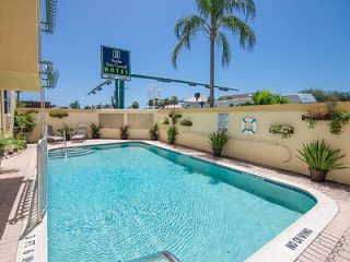 Outdoor swimming pool at a hotel with palm trees and a blue sky, surrounded by a cream-colored wall, and a hotel sign visible in the background.