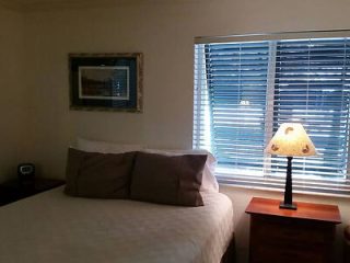 A neatly made bed with pillows beside a nightstand with a lamp, under a window with partially open blinds in a warmly lit room.