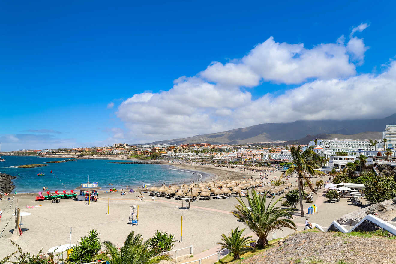 A vibrant beach scene with visitors enjoying the sandy shore, parasols, and a panoramic view of a bustling resort town of Costa Adeje