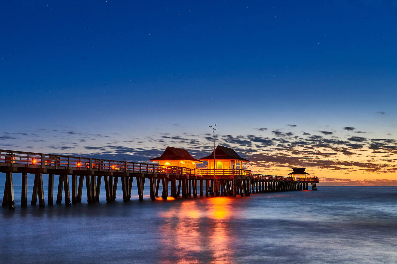 Twilight descends on a wooden pier extending into the ocean, with glowing lights illuminating the structure against a dramatic sky of blue hues and scattered clouds