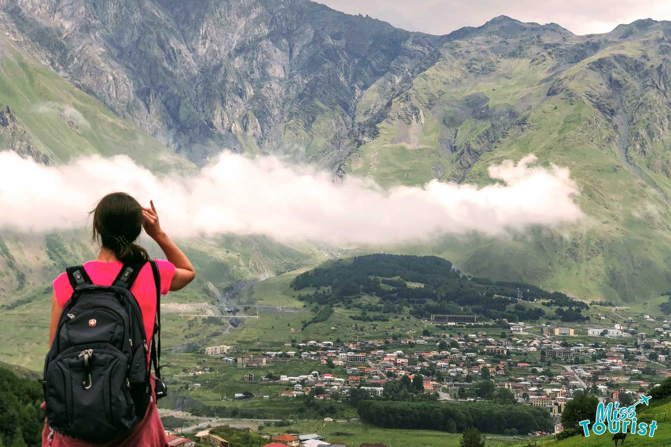 The writer of the post with a pink top and a black backpack gestures towards a sweeping view of a valley with low-hanging clouds and green mountains, with the quaint buildings of a village nestled below