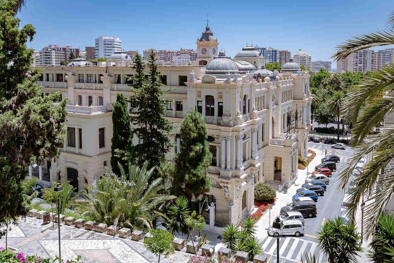 Elegant historic building in Malaga with a facade of ornate architecture, surrounded by lush greenery and parked cars on a sunny day