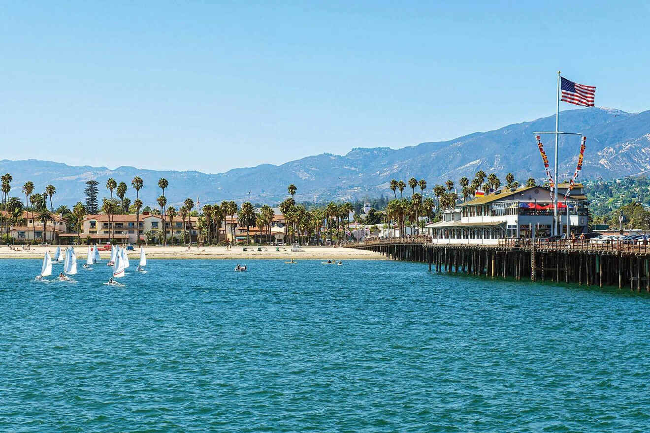 "Santa Barbara pier extending into the sparkling ocean, with small sailboats navigating the waters and palm trees framing the mountainous horizon