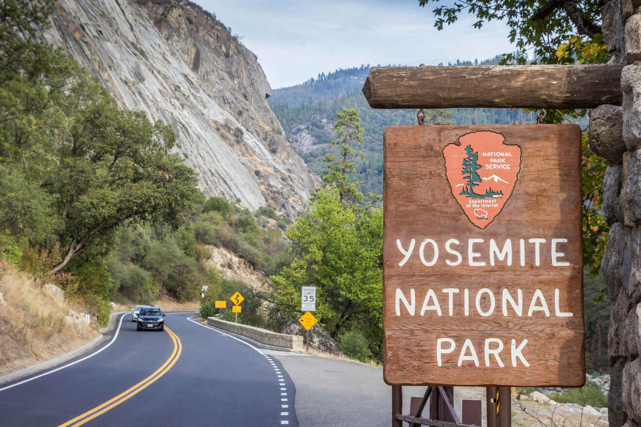 Entrance sign of Yosemite National Park on a scenic road flanked by forested slopes and a clear blue sky.
