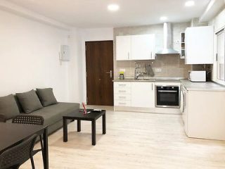 Modern, minimalist apartment interior with a combined living room and kitchen area featuring a grey couch, black coffee table, and white kitchen appliances.