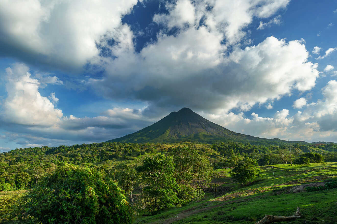 A panoramic view of a green landscape with a prominent volcano, surrounded by scattered trees and a partly cloudy sky.