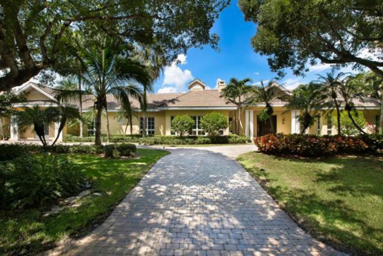 Elegant single-story house with a yellow facade and tiled roof, featuring a cobblestone driveway surrounded by lush palm trees and manicured landscaping under a bright blue sky