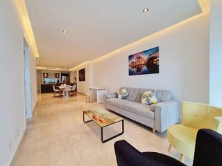 Modern living room with sleek furniture, a large wall-mounted painting, and an open-plan layout leading to a dining area under soft ceiling lights