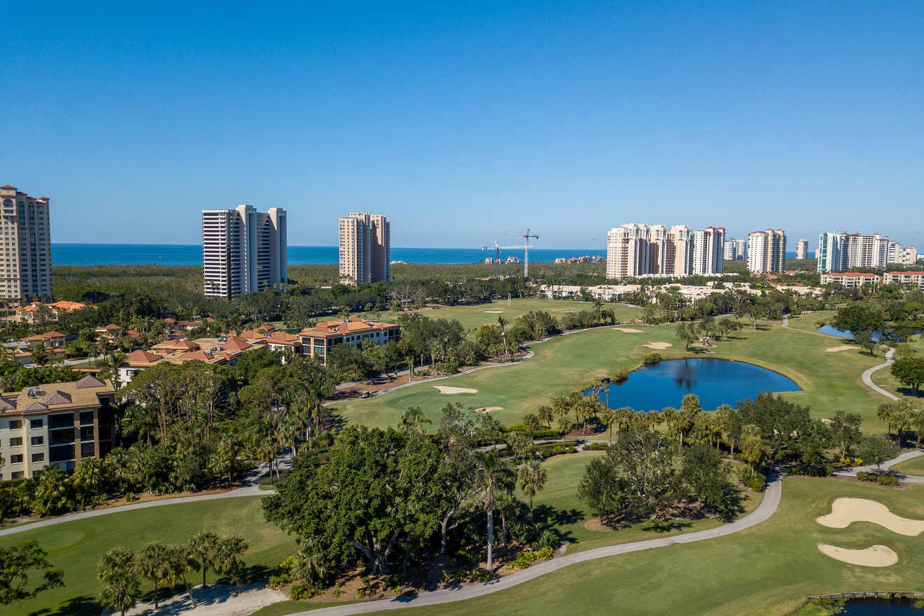 Aerial view of a lush golf course with winding paths and sand bunkers, flanked by clusters of high-rise buildings with ocean views in the background under a clear blue sky