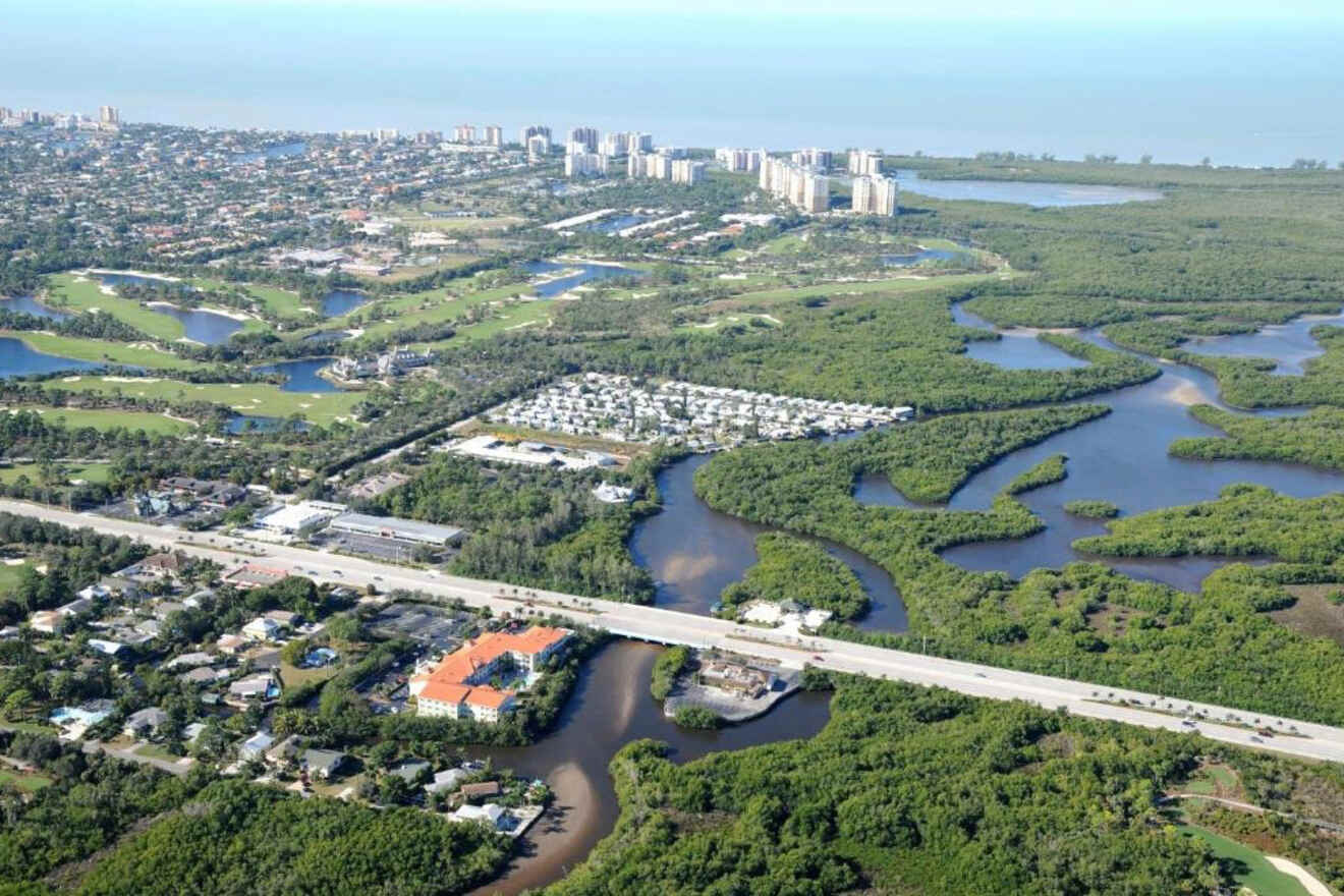 Expansive aerial view showcasing a mix of residential areas, golf courses, and natural mangrove estuaries with snaking waterways, leading towards a skyline of high-rise buildings by the coast